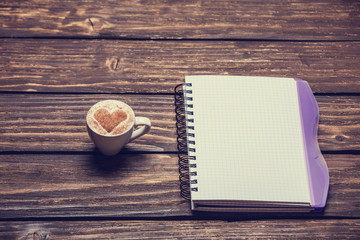 Cup of coffee with shape heart and notebook on wooden table.