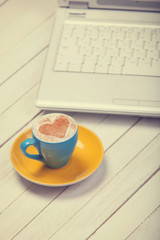 Cup of coffee with heart shape and notbook on wooden table.