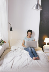 Casual young woman sitting on bed and using laptop at home.