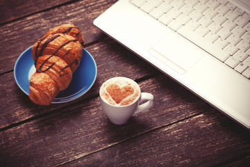 Croissant and cup of coffee with laptop on wooden table.