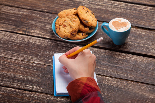 Female hand writing something in note near cookie and cup of cof