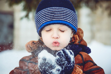 Little boy blows snow with mittens on bokeh background of snowfl