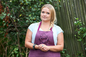 Lush woman in apron with folding hands standing near rural fence