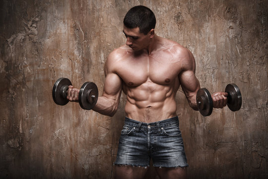 Naked muscular man working out with dumbbells on wall background