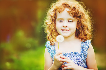 Beautiful little curly girl blowing dandelion, horizontal shot.