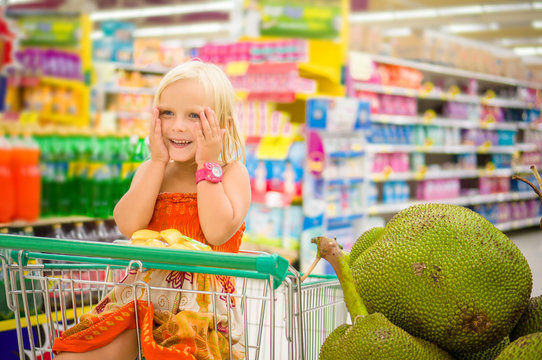 Adorable girl in shopping cart looks at giant jack fruits on box