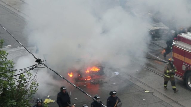 Riot officers and fireman walking around car fire with smoke