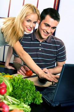 Young happy couple cooking at kitchen with laptop