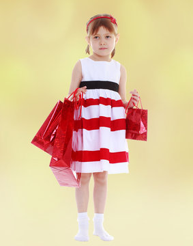 Girl in a striped dress holding red paper shopping bags.