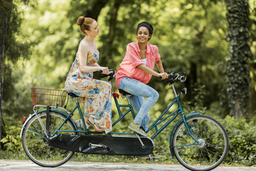 Young women riding on the tandem bicycle - obrazy, fototapety, plakaty