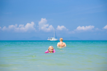 Young mother and daughter swim in clear tranquil ocean with yach