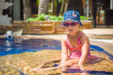 Adorable girl with pink sunglasses and blue hat sit in pool on s