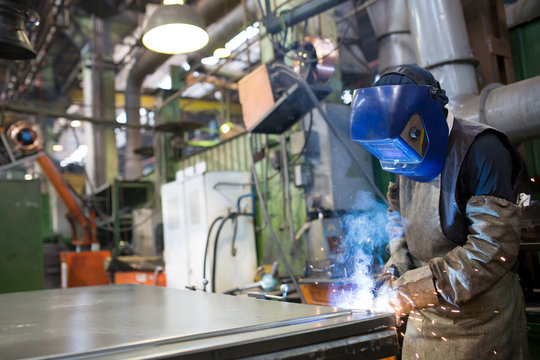 Worker welding metal at factory workshop with flying sparks