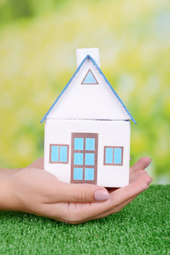 Woman hand holding small house on grass on bright background