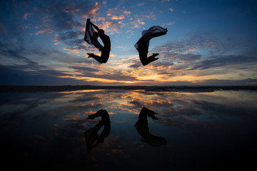 two silhouetted girls jumping in sunset on beach with reflection - 70193421