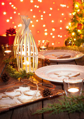 Christmas table setting. Elegant white plates with cookies, natural pine tree branch, pinecones, candles, lights and gifts on vintage planked wood. Rustic style decorations on red bokeh background.