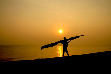 Silhouettes of a man walking carrying a timber beside the sea.