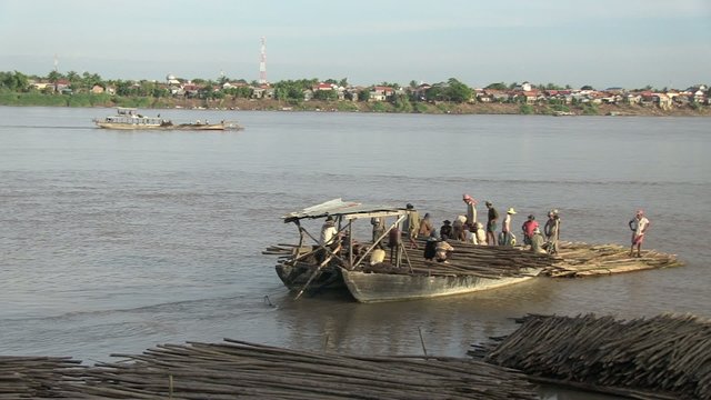 Barge drawing away from the riverbank transporting workers and bamboo poles laid width-wise across/towing a heap of bamboo from the bow;