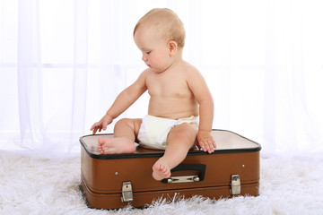 Cute baby boy sitting on suitcase in room