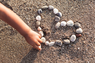 Baby's hand making heart of marine shingles on the sand.