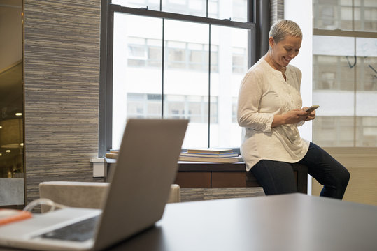 Office life. A woman seated on the edge of her desk using a digital tablet. 