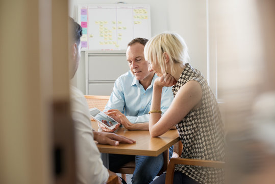 Three business colleagues in an office talking around a table and looking at a digital tablet. 