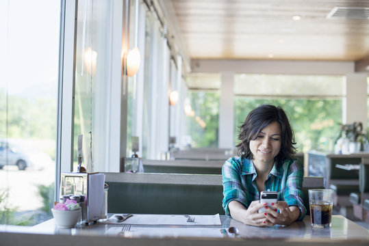 A woman in a checked shirt sitting at a table, laughing and looking at her smart phone.