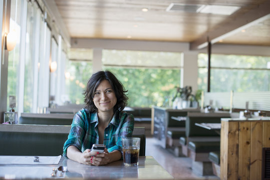 A woman in a checked shirt sitting at a table, laughing and looking at her smart phone.