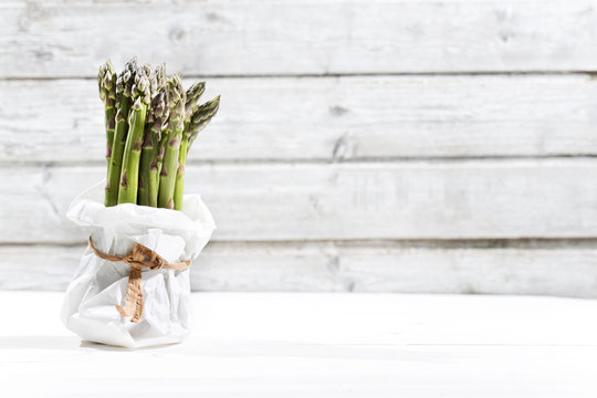 Bunch of green asparagus standing in white paper bag in front of white wood