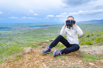 girl looks with binoculars sitting on mountain rock