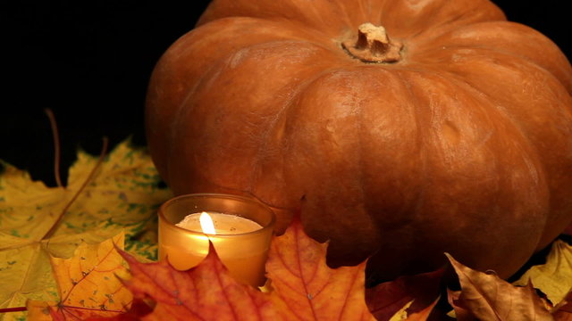 The candle burns before pumpkin and maple leaves on dark backgro