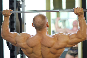 Back of bodybuilder raising barbell in gym hall before mirror