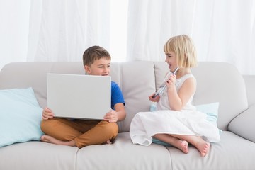 Siblings using a laptop and a tablet sitting on a couch