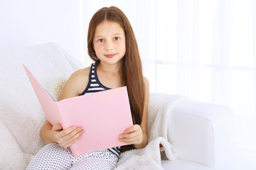 Beautiful little girl reading book on sofa in room