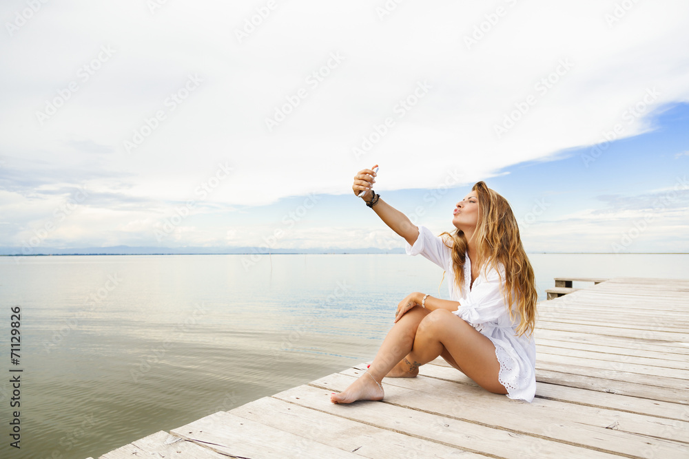 Wall mural beautiful young woman sitting on a pier and taking a self portra