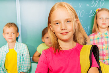 Girl wears bag near blackboard, pupils behind her