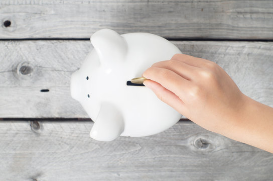 Asian Boy Hand Dropping A Coin Into White Piggy Bank  Against Wo