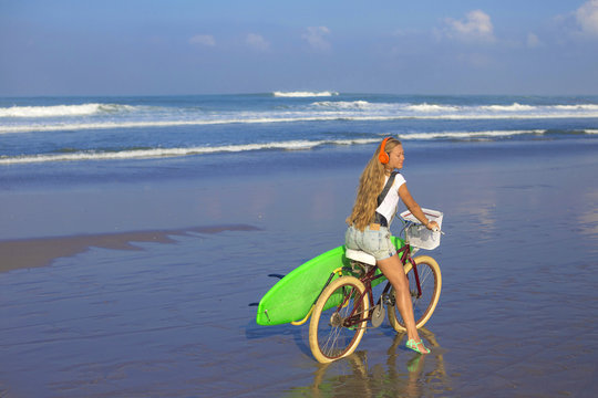 Young girl with surfboard and bicycle on the beach.