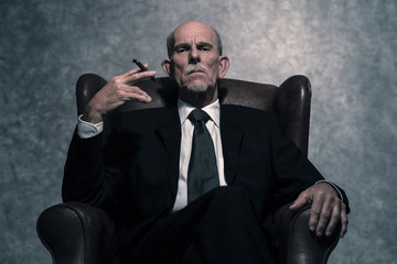Cigar smoking senior businessman with gray beard wearing dark su