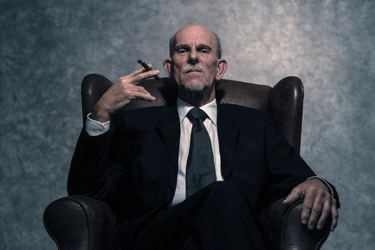 Cigar smoking senior businessman with gray beard wearing dark su