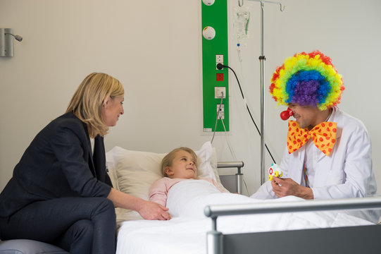 Male doctor wearing clown costume making girl patient smile in hospital bed