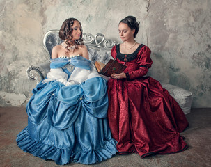 Naklejka premium Two beautiful women in medieval dresses on the sofa reading book