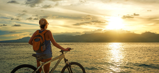Young woman with backpack standing on the shore near his bike.