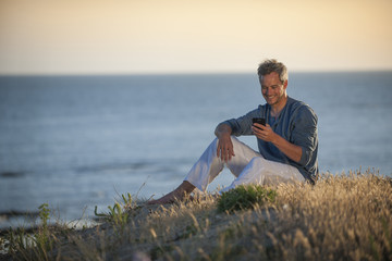 handsome man sitting on the beach at sunset and writing a sms on