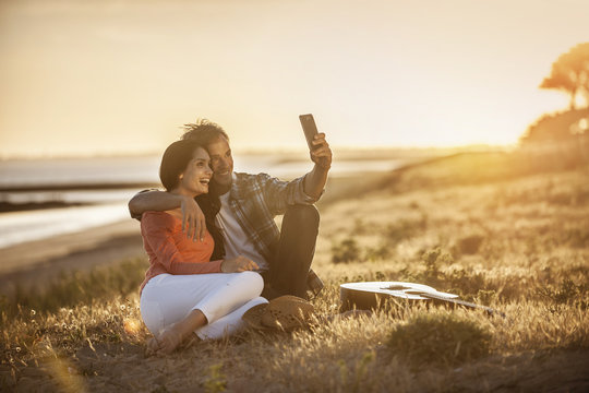 Romantic couple sitting on the beach at sunset and taking a self