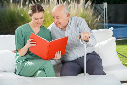 Senior Man Reading Book With Female Caretaker On Couch
