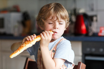 Little boy eating long loaf bread or baguette in kitchen.