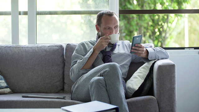 Businessman with smartphone drinking coffee on sofa at home