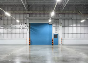 Selbstklebende Tapeten Industriegebäude Rolltor oder Rollladen in Fabrik, Lager oder Industriegebäude. Modernes Innendesign mit poliertem Betonboden und leerem Raum für Produktpräsentation oder Industriehintergrund.