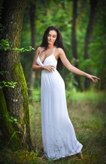 Lovely young lady wearing an elegant long white dress in forest